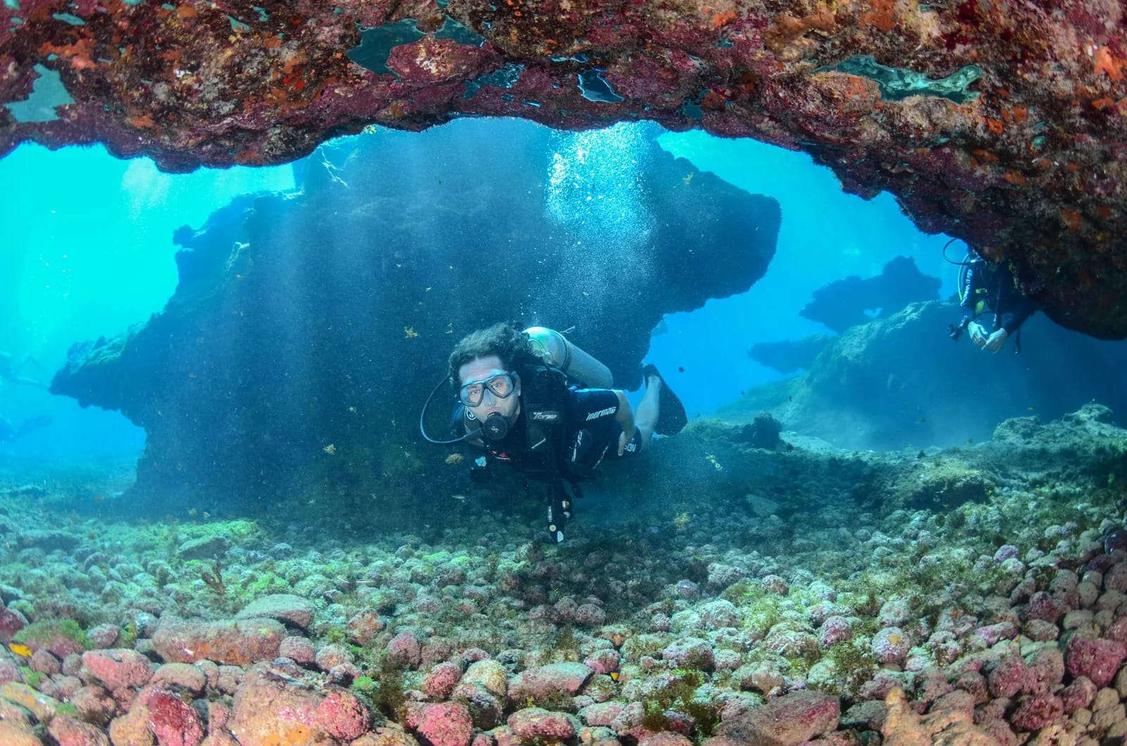 Plongeur sous une arche rocheuse sous-marine, lumière qui filtre dans une eau turquoise