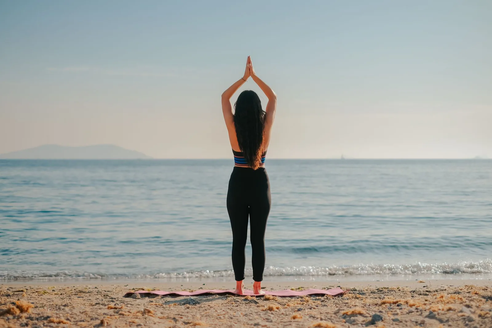 Femme en posture de yoga, mains jointes au-dessus de la tête, face à la mer sur une plage