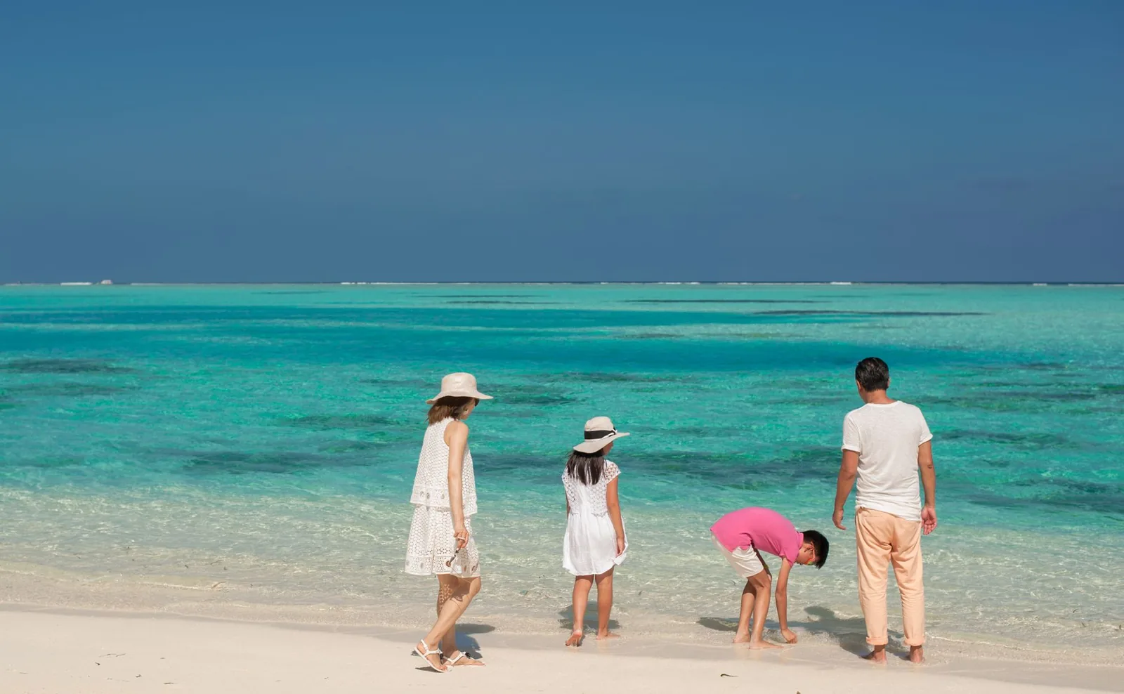 Famille avec deux enfants les pieds dans l'eau turquoise d'un lagon, sur une plage de sable blanc