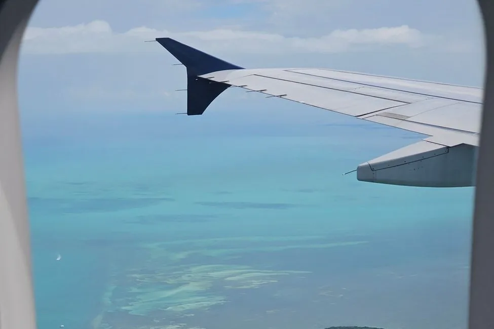 Vue par le hublot d'un avion : aile et mer turquoise bordée de hauts-fonds clairs